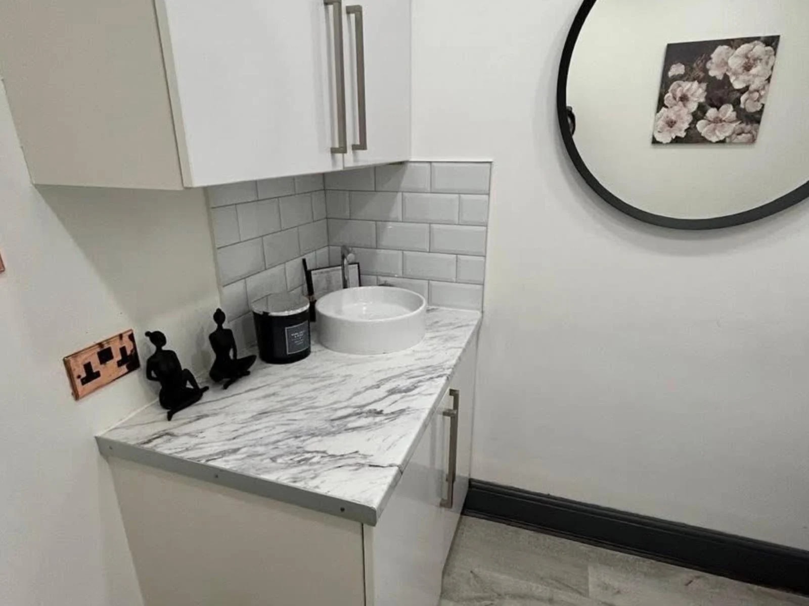 Treatment-room sink area with white cabinetry, marble-effect worktop and circular mirror